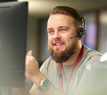 a call centre phone operative in his mid 30s chats on the phone at his desk . He is explaining something to the person on the phone in a friendly manner . behind him a defocussed office interior can be seen . This could be a call centre or an office worker chatting to a customer .